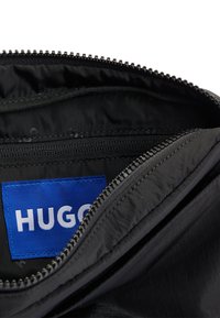 Bolsa con cremallera negra de exterior texturizado, con una etiqueta de tela azul que dice "HUGO" y un bolsillo interno con cremallera.