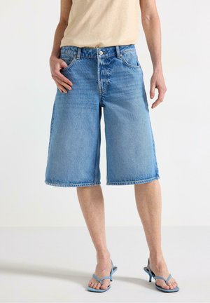 Person iført løse, knælange lyseblå denimshorts, beige top og lyseblå hælede sandaler, stående med én hånd i lommen.