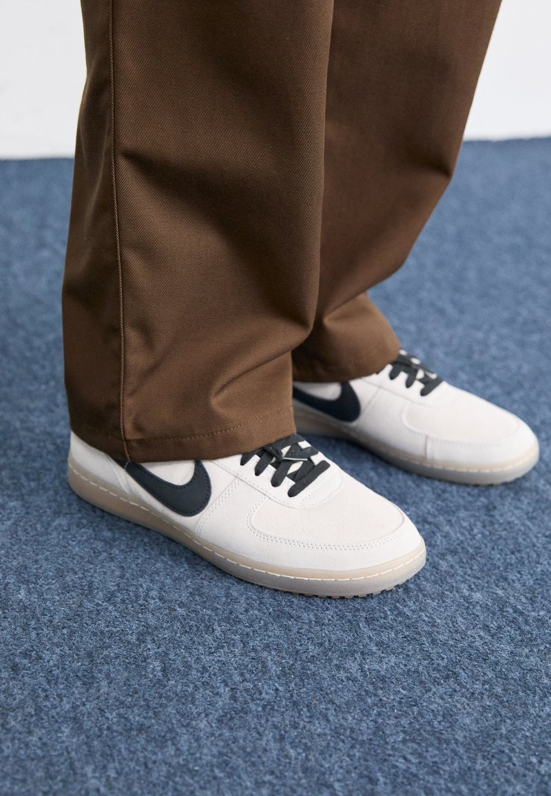 Sneaker bianche con accenti neri, realizzate in tessuto liscio, caratterizzate da un design a profilo basso e punta tonda. Pantaloni marroni che coprono parzialmente le scarpe.