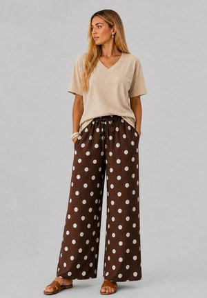 Femme debout, les mains dans les poches, portant un t-shirt beige à col en V et un pantalon marron à jambes larges avec de grands pois blancs et des sandales marron.