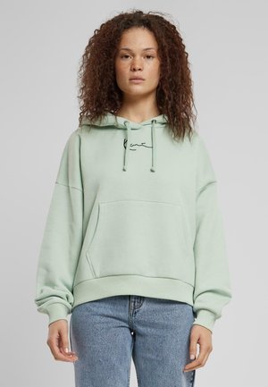 Menta zöld, oversized kapucnis pulóver, elülső zsebbel, húzózsinórral és hímzett részletekkel. Világoskék farmernadrággal párosítva.