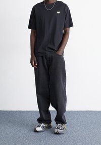 Camiseta de algodón negra con un logo dorado, combinada con pantalones vaqueros de mezclilla oscuros y holgados, y zapatillas grises con detalles amarillos.