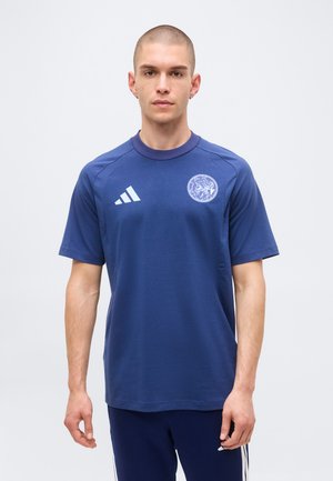 Jonge man met geschoren hoofd die een blauw Adidas sportshirt en bijpassende broek draagt tegen een effen lichte achtergrond.