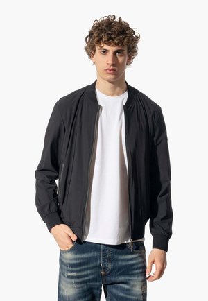 Giubbotto bomber nero con polsini e vita elasticizzati, tessuto leggero, zip frontale, indossato sopra una maglietta bianca e jeans blu strappati.