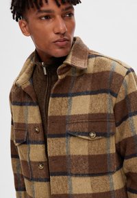 Brunternet plaid uldjakke med blå og beige accenter, der har to forlommer og en krave, båret over en struktureret brun top.