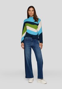 Frau in einem blau-grün-weißen gestreiften Pullover, weit geschnittenen blauen Jeans und weißen Sneakers, die vor einem schlichten grauen Hintergrund steht.