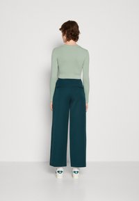 Person med kort krøllet hår iført lysegrøn langærmet top, mørke vide bukser og hvide sneakers, står mod en ensfarvet baggrund.