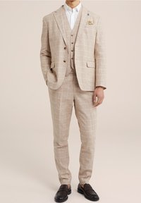 Světle béžový kostkovaný oblek s upraveným střihem, obsahující sako na jedno knoflíkové zapínání, klopové límce a odpovídající kalhoty. Černé mokasíny dotvářejí vzhled.