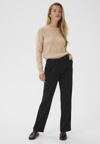 Femme se tenant debout avec une main dans la poche, portant un pull beige côtelé, un pantalon large noir avec des boutons et des mocassins bordeaux sur fond blanc.