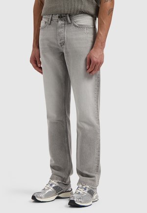 Mann trägt hellgraue Jeans mit geradem Bein und silberne Sportschuhe, steht vor einem schlichten hellen Hintergrund.