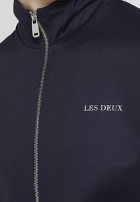 Námořnická zipová bunda s vysokým límcem, stříbrným zipem a bílým textem "LES DEUX" na hrudi, vyrobená z hladké, elastické látky.