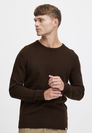 Ung mand med kort hår iført mørkebrun sweater og beige bukser, kigger mod sin venstre side med hænderne foldet foran sig.