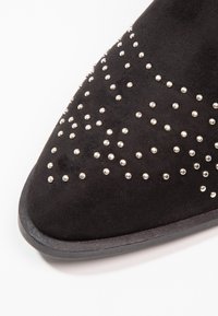 Zapato de ante negro con punta afilada, adornado con tachuelas plateadas. Presenta una suela lisa con textura mínima, que resalta el diseño con tachuelas.