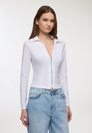 Camicia bianca con zip, colletto, maniche lunghe e silhouette aderente, abbinata a jeans in denim blu chiaro. Tessuto liscio, design minimalista.