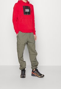 Sudadera con capucha roja con logo negro, pantalones cargo oliva y zapatillas deportivas negras con detalles en naranja. La sudadera tiene un bolsillo frontal y un cordón ajustable.