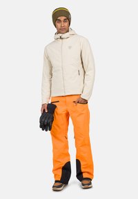 Giacca isolante beige chiaro con cappuccio, abbinata a pantaloni impermeabili arancioni brillanti e guanti neri. Caratterizzata da tasche con zip e dettagli a contrasto.