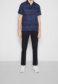 Korte mouwen blauw en zwart geruite overhemden, met knopen, gedragen met zwarte broek en witte sneakers, staand tegen een effen achtergrond.