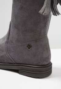 Bottine en suède grise avec semelle en caoutchouc noire et petit logo en métal rond près du talon, présentant un détail de pompon en suède en haut.