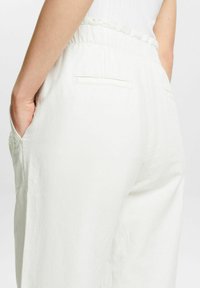 Pantalon en lin blanc avec une coupe décontractée, taille élastique à détail volanté et poches latérales. Le dos comporte deux poches passepoilées.