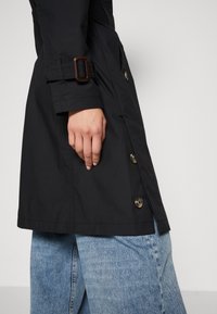 Main reposant le long du corps, portant un manteau noir boutonné avec une sangle à boucle sur la manche et un jean en denim bleu clair.