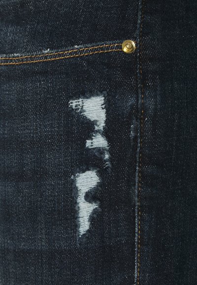 Jeans in denim blu scuro con una sezione strappata e bordi sfrangiati, con tessuto bianco sottostante. Hardware dorato e cuciture arancioni visibili.