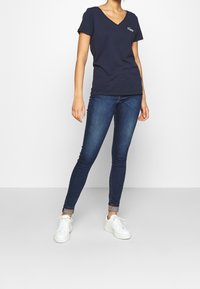 Mörkblå bomulls t-shirt med V-ringning och korta ärmar, kombinerad med mörkblå smala jeans och vita sneakers. Har en liten logotyp på bröstet.