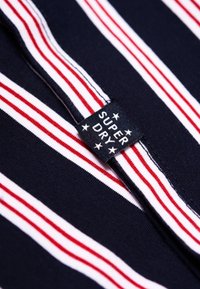 Marine stof met horizontale rode en witte strepen; heeft een zwart label met de tekst "SUPER DRY" en sterren. Getextureerd brei materiaal.
