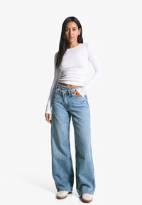 Femme portant un top court à manches longues blanc, un jean large bleu clair et des baskets blanches, se tenant devant un fond blanc uni.