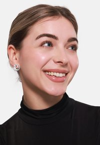 Boucles d'oreilles en argent évoquant des formes circulaires entrelacées avec une finition polie, portées par une personne à la peau claire et aux cheveux foncés.