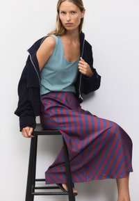 Tumši zila jaka, gaiši zils tops bez piedurknēm un sarkani tumši zils svītrains garais svārks. Gluda auduma, brīva piegrieztne, un ikdienišķa sēdēšanas poza uz krēsla.