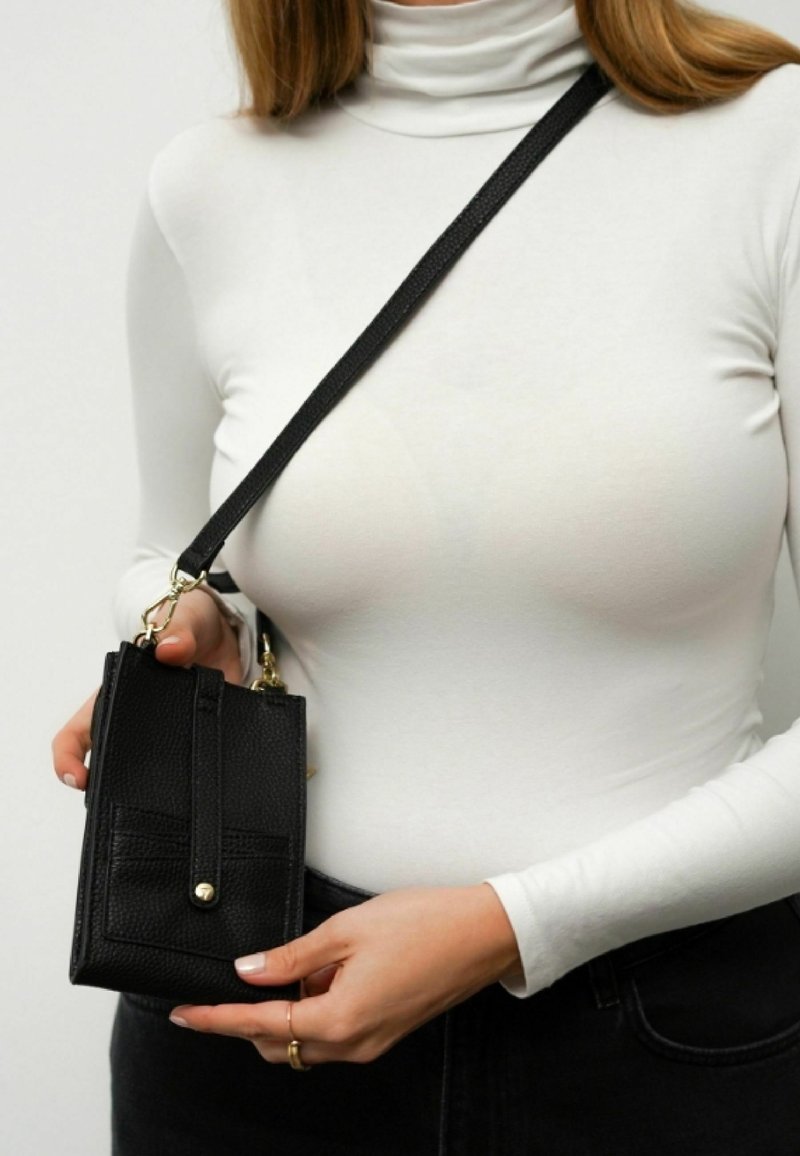 Schwarze, strukturierte Umhängetasche mit einer Fronttasche und goldenen Beschlägen, gehalten von einer Person in einem weißen Rollkragenoberteil.