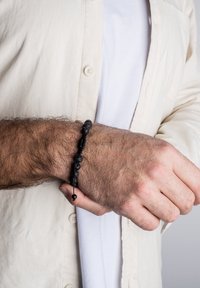 Brăţară țesută neagră cu mărgele gri texturate, cord ajustabil cu închidere prin mărgeluță, purtată pe un încheietura mâinii, pe un fond de cămașă de culoare deschisă.