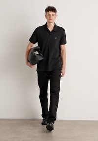 Zwarte polo, slim-fit zwarte broek, zwarte schoenen, met een grote zwarte tas. Eenvoudig ontwerp met een subtiele logo op het shirt.