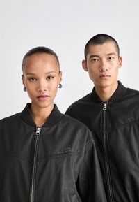 To modeller iført sorte zip-up jakker med "HUGO" præget på brystet, der står imod en ensfarvet lys baggrund.