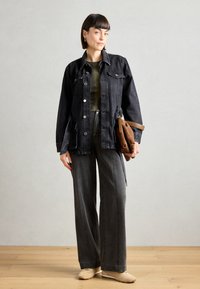 Giacca di jeans nera con bottoni argentati, indossata sopra una maglietta verde oliva, abbinata a pantaloni larghi grigio scuro e scarpe beige. Accessorio in borsa marrone.
