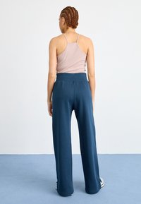 Kvinna med flätat hår står med ryggen mot kameran, iklädd en ljus mauve linne och lösa marinblå byxor, på en blå golv och vit vägg bakom.