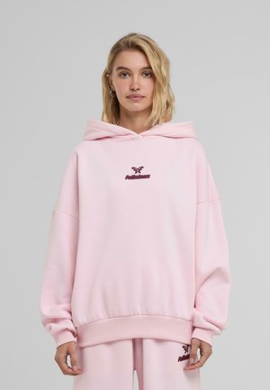 Felpa oversize rosa realizzata in morbido tessuto, presenta un cappuccio con coulisse e un logo bordeaux ricamato sul petto. Polsini e orlo a costine.
