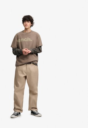 Młody mężczyzna z kręconymi włosami stoi, ubrany w brązowy t-shirt Bench na koszulce z długim rękawem, beżowe spodnie i czarne trampki.
