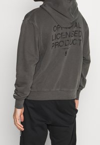 Grau Hoodie aus weichem Material, mit großem schwarzem "OFFIZIELL LIZENZIERTES PRODUKT"-Text auf dem Rücken und einer Kapuze mit Kordelzug.