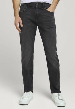 Uomo che indossa jeans neri slim fit e sneakers bianche, con una mano in tasca e camicia bianca parzialmente visibile.