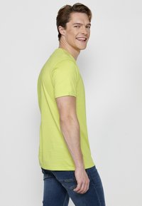 Tricou din bumbac de culoare verde lime, cu mâneci scurte, guler rotund și o textură netedă. Purtați cu blugi albaștri, văzut din lateral.