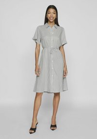 Páskované košilové šaty v šedé a bílé barvě, s krátkými rukávy, staženým pasem a sukní po kolena, v kombinaci s černými sandály na otevřené špičce.