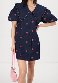 Femme portant une robe boutonnée bleu marine avec des manches bouffantes et des motifs de cœurs rouges, tenant un petit sac à main rose dans une main.