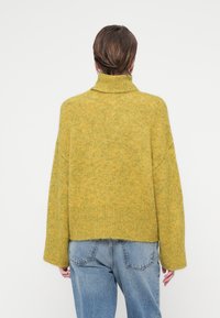 Hořčicový svetr s vysokým límcem, texturovaným pletením a oversized střihem. Disponuje spadnutými rameny a žebrovanými manžetami, v kombinaci s modrými džíny.
