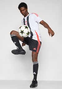 Hvid og sort fodboldtrøje med røde accenter, sorte shorts, sorte støvler og stribede sokker; spiller der balancerer en fodbold på sit knæ.