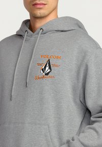 Grijze sweatshirt met capuchon, voorzien van een vooraanstaande grafische logo in oranje en zwart. Inclusief trekkoorden en een kangoeroezak.