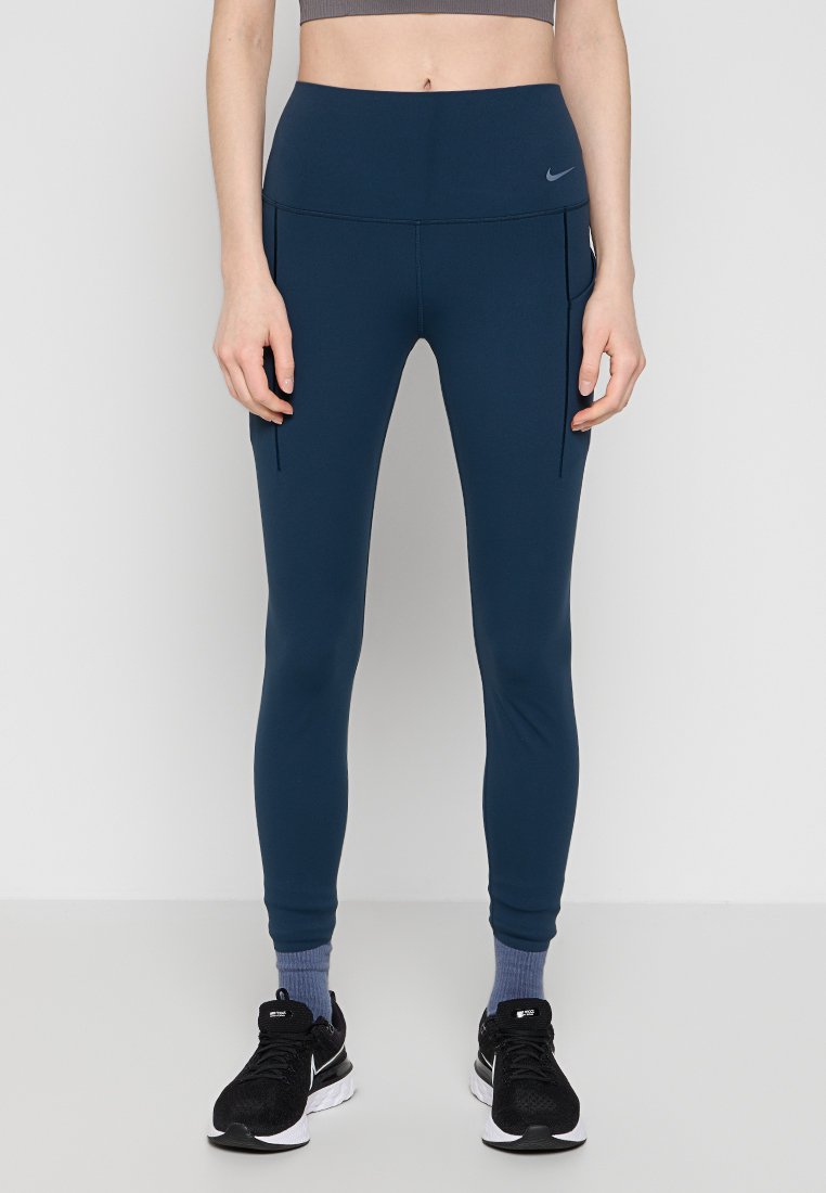 Persona con leggings de talle alto azul marino con bolsillos laterales, calcetines de tobillo grises y zapatillas de correr negras de Nike, de pie contra un fondo blanco.