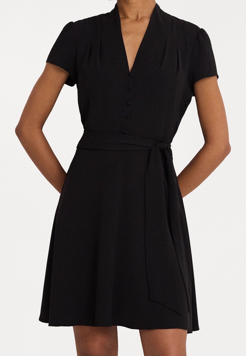 Svart klänning med korta puffärmar, djup V-ringning, knappstängning och knytning i midjan. Tyget är slätt och något strukturerat.