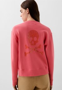 Korálový sweatshirt s žebrovanými manžetami a lemem, na zadní straně s třpytivým designem lebky a zkřížených kostí. Měkká, středně těžká látka.