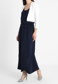 Granatowa sukienka maxi z marszczonym pasem, zestawiona z krótką białą kurtką. Modelka ma na sobie jasnoszare szpilki. Gładki materiał, elegancki krój.